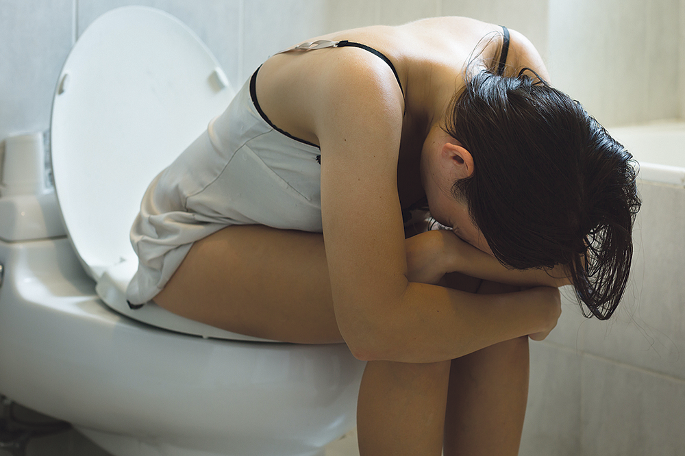 Eine Frau sitzt vor Schmerzen vorne über gebeugt auf der Toilette.