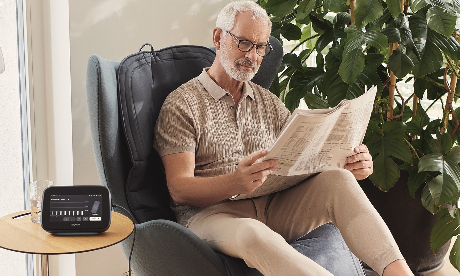 Älterer Mann sitzt mit Zeitung in der Hand im Sessel während er B. BODY Evo von BEMER nutzt