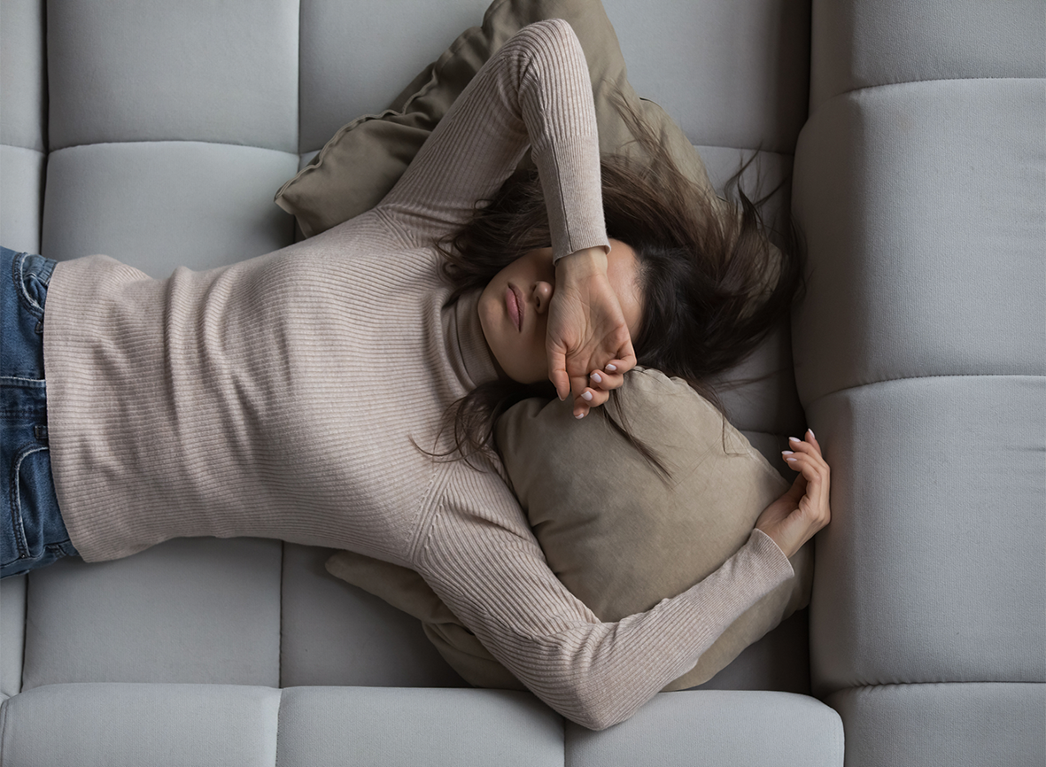 Frau liegt auf dem Rücken auf einem grauen Sofa mit dem Arm über ihrem Gesicht.