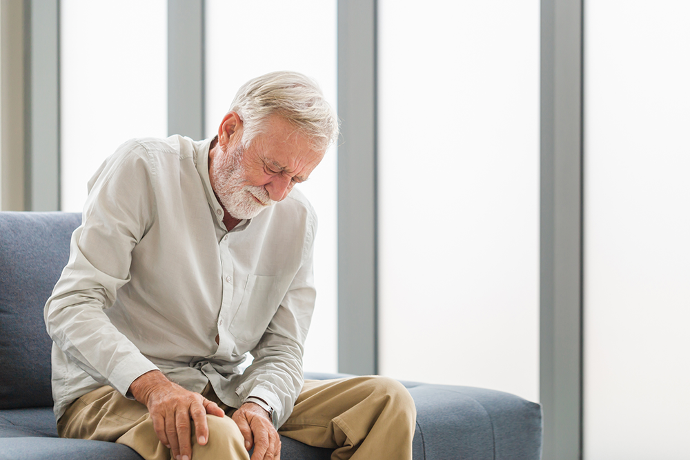 Ein älterer Herr hält sich mit schmerzverzerrtem Gesicht auf einer Couch sitzend das Knie.