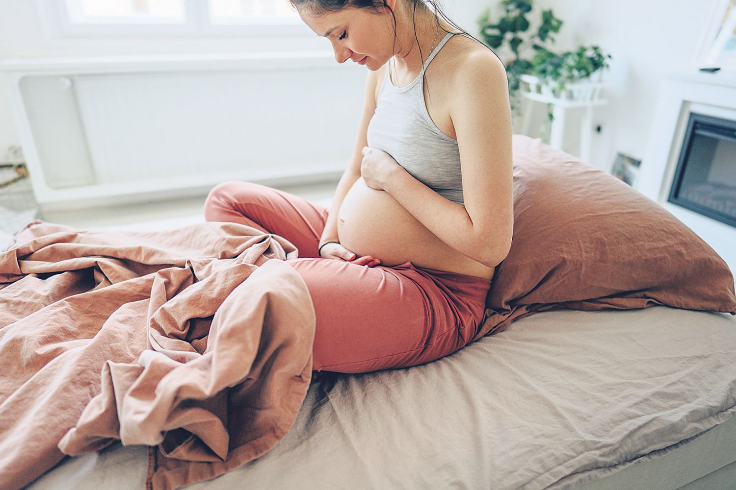 Eine schwangere Frau sitzt im Bett und hält ihren Bauch, während sie ihn anblickt. 